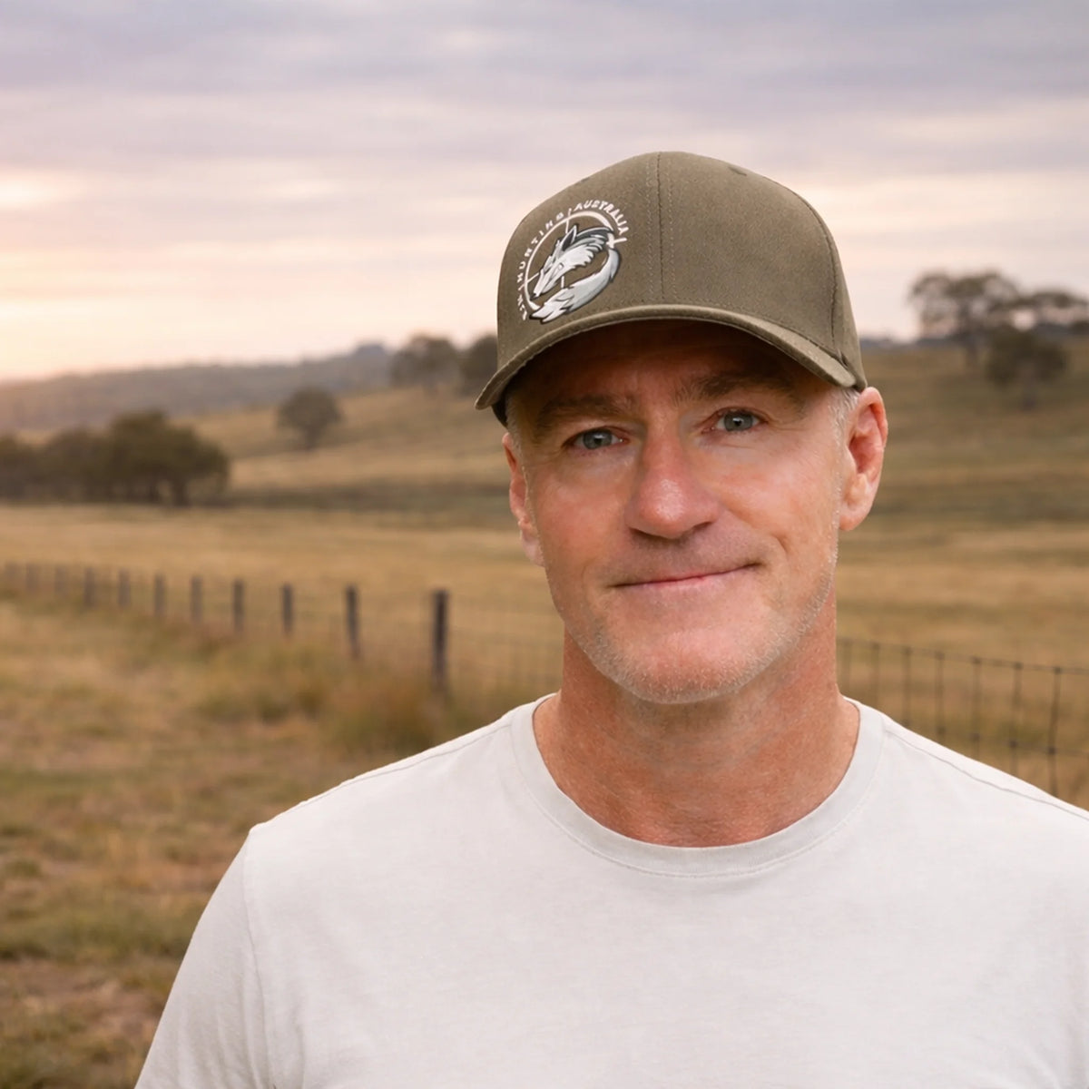 Man in a white t-shirt wearing an olive RJM Hunting Australia cap on a country property